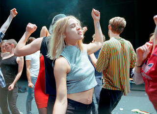 Eine Gruppe von Tänzerinnen auf der Bühne; im Zentrum tanzt eine junge Frau mit blonden Haaren mit geschlossenen Augen.