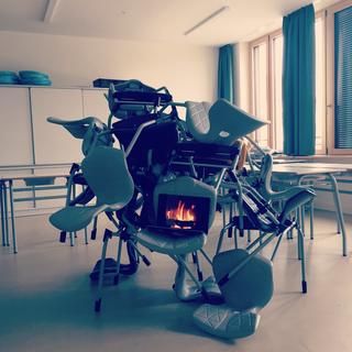 Stühle sind in einem Klassenzimmer zum BErg gestapelt. Auf einem Laptop glüht ein Lagerfeuer.
