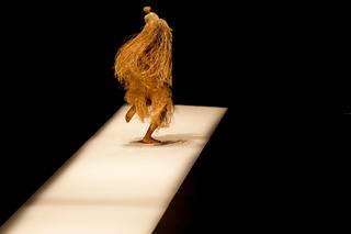 Ein Performer in einem fransigen Ganzkörperkostüm tanzt auf einem beleuchteten Laufsteg.