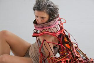 Practicing Empathy #3 Eine Performer*in mit einem futuristischen Kostüm schaut auf dem Boden sitzend nach links