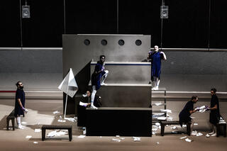 Eine graue Bühne. Eine Treppe mit Gucklöchern. Performer*innen in blauer Kleidung.