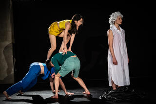 Drei Performer*innen bilden durch akrobatische Bewegungen eine Pyramide. Eine weitere Person in weiß gekleidet steht neben der Gruppe.