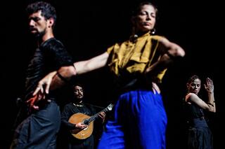 Auf der Bühne sind drei Performende zu sehen, die den Fado Batido aufführen, eine Art des Stepptanzes. Im Hintergrund spielt eine Person eine Art Gitarre als musikalische Unterstützung.