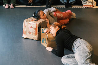 Zwei weiblich gelesene Tänzer*innen liegen auf der Seite auf dem Boden. Ihre Köpfe haben Sie wie zum Schlafen auf Kartons abgelegt.