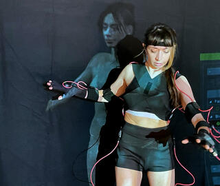 Eine Performerin trägt einen Motion-Capture-Anzug, mit dem ein Avatar generiert wird, der im Hintergrund zu sehen ist.