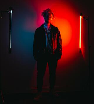 Felix Waltz von LA CRUSH Künstlerisches Foto von Felix Waltz (LA CRUSH Electro Musik) im Halbdunklen, beleuchtet von Lichtröhren, wobei eine davon rot auf ihn strahlt.
