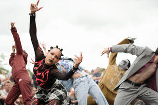 Gruppe von Tänzer*innen strecken sich in verschiene Richtungen in verschiedenen Outfits. Vorne ist eine Tänzerin mit roten Flammenshirt und Schottenrock zu sehen