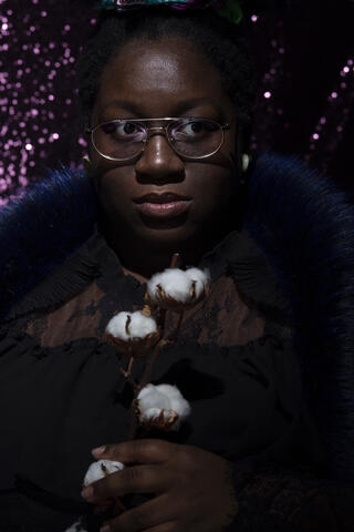 Ein Portraitfoto der DJane T/Error. Sie hält einen Zweig mit Baumwollblüten in der Hand. Sie trägt eine Brille und ihr Blick ist zur Seite gerichtet.