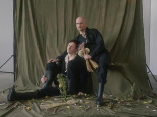 it (still) is what it is Zwei Performer sitzen schwarzer Kleidung und schwarzen Schuhen in einem Fotoshooting-Setting. Dunkelgrün-Graue Vorhang im Hintergrund. Der Hintere sitzt auf einer Erhöhung, der Vordere sitzt auf dem Boden und lehnt sich an den Hinteren. Auf dem Boden liegen vertrocknete Blumen.
