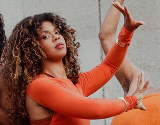 Die Tänzerin und Choreografin ZOE ist im Seitenprofil zu sehen. Sie trägt ein orangenes Kleid mit langen Ärmeln. Ihre Arme hat sie zu ihrer linken Seite auf Brusthöhe ausgestreckt, ihre Finger sind gerade und angespannt. Sie schaut mit leicht gehobenem Kinn Richtung Betrachter*in.