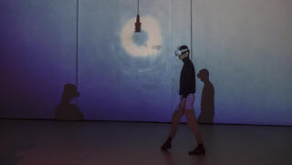 Performer mit VR-Brille läuft von rechts nach links über die blau beleuchteten Bühne. Im Zentrum eine Lampe, die einen kreisförmigen Lichtring wie ein Heiligenschein an die Wand hinter sich wirft.