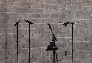 Fünf Mikrofonständer stehen vor einer grauen Backsteinwand.