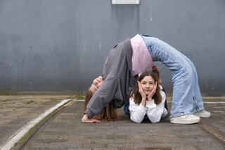 Zwei Jugendliche vor eine grauen Wand und auf einem grauen Backsteinboden. Eine Jugendliche meine eine Brücke rückwärts über die andere Jugendliche die dazwischen liegt und den Kopf in die Hände legt.