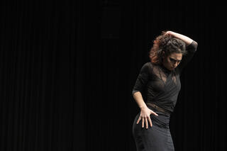 Das Bild hat einen schwarzen Hintergrund. Rechts ist eine Person abgebildet, ebenfalls in schwarz gekleidet und in einer szenischen, tanzenden Bewegung. Eine Hand liegt auf ihrer Hüfte, die andere an ihrem Kopf, es scheint als halte die ihre Haare fest. Die Person blickt in die rechte Ecke des Bildes auf den Boden. 