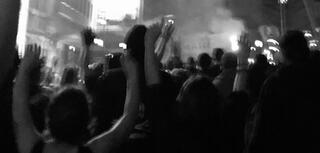 Eine große Gruppe von Personen ist von hinten zu sehen. Sie feiern gemeinsam, einige von ihnen halten ihre Arme in die Luft. Das Foto ist schwarz-weiß und unscharf.