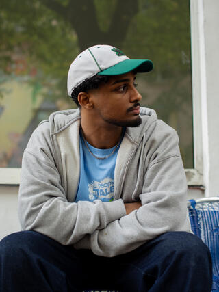 Auf dem Bild ist eine Person zusehen die sitzt. Sie stützt ihre Arme verschränkt auf ihren Beinen ab. Außerdem schaut die Person zur Seite.