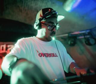 Auf dem Bild ist eine Person zusehen die Kopfhörer trägt. Sie hat beide Arme auf einem Pult und schaut nach unten. das Bild ist von grünem Licht erleuchtet. 