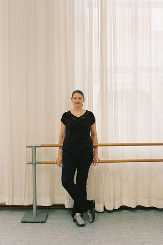 Das Bild bildet eine stehende Person in schwarz gekleidet ab. Sie lehnt an einer Ballettstange und schaut in die Kamera. Vor ihr ein grauer Boden und hinter ihr weiße Vorhänge von Licht durchflutet.