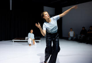 Auf einer Bühne mit weißem Boden steht im Vordergrund eine Person, eine weitere sitzt im Hintergrund vor einem flachen Podest. Beide Personen tragen Hosen und T-Shirts. Die Person im Vordergrund nimmt eine Position wie beim Kugelstoßen ein: Ihren linken Arm streckt sie zur Seite, leicht nach oben gerichtet, den anderen Arm hält sie angewinkelt auf Oberkörperhöhe. Beide tragen ein Headsetmikrofon.