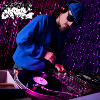 Das Bild zeigt eine Person die an einem DJ Pult steht. Sie trägt blaue Klamotten. Das Bild ist lila-pink beleuchtet.