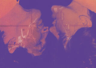 Das Bild zeigt zwei Personen. Diese beiden Personen stehen auf dem Kopf. Es ist nur deren Profil zusehen. Die beiden sind nah aneinander und schauen zu einander. Auf ihnen ist eine Projektion zusehen, von einer weiteren Person. Das Bild ist lila - orange eingefärbt.