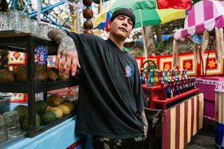 Das Bild zeigt eine Person die ihren einen Arm geknickt nach oben hält. Im Hintergrund sind bunte Stände mit Obst und Gläsern zusehen.