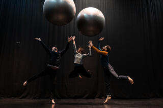 Zwei große silberne Bälle fliegen in die Luft auf einer schwarzen Bühne. Darunter drei Performer*innen die nach hinen springen und die Hände in der Luft haben. Sie sind barfüßig und haben Alltagskleidung an.