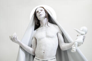 Auf dem Bild ist eine Person abgebildet, die weiß angemalt ist. Der Oberkörper ist nackt, ein Umhang liegt über ihrem Kopf und sie trägt ein Baby in der linken Hand. Als würde sie beten schaut sie hoch und hält die Arme nach oben, Handflächen gen Himmel.