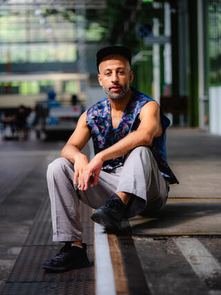 Ein Portraitfoto des Tänzers Rymon Zacharei aka Rayboom. Er sitzt im urbanen Raum auf einer Stufe. Seine Hände liegen lässig auf seinen Knien und er trägt ein Streetstyle-Outfit mit grauer Hose, einem kurzärmeligen Oberteil und einer Kappe.
