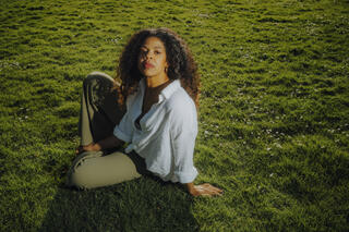 Ein Foto der Performance-Künstlerin und Kuratorin ZOE (Zoe-Marie Buchholz). Sie sitzt mit angewinkeltem Knie auf einer Wiese. Sie trägt eine hellgrüne Hose und ein weißes Musselinhemd. Mit neutralem Blick schaut sie leicht an der Kamera vorbei.