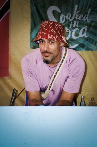 Eine männlich gelesene Person mit einem Schnurr- und Kinnbart trägt ein rosa Shirt und einen roten, gemusterten Bucket Hat. Sie steht hinter einer Theke. Die Person schaut in die Kamera und lächelt. Hinter der Theke sind Glasflaschen zu erkennen.