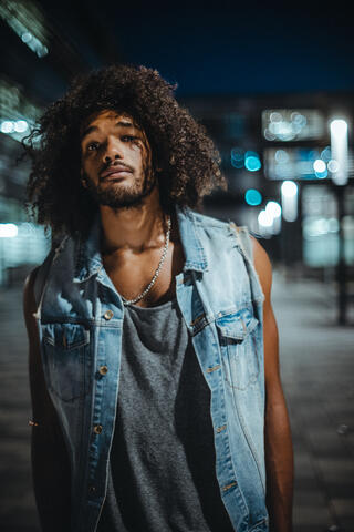 Ein Portraitfoto des Tänzers Paul Davis Newgate in einer Nachtszenerie. Im Hintergrund sind verschowmmene Gebäude zu erkennen. Er trägt eine Jeansweste und ein graues Tanktop.