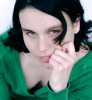 Ein Portraitfoto der Künstlerin Gaia Pellegrini, von oben fotografiert. Sie hat schwarze schulterlange Haare und eine Strähne hängt in ihr Gesicht. Sie stützt ihr Kinn auf ihrer Hand ab und schaut nach oben in die Kamera. Sie trägt einen grünen Cardigan.
