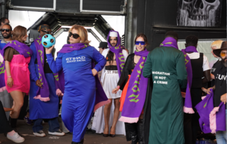  Gruppe in verschiedenen Outfits, mehrere tragen Schals mit Schrift, eine Jacke zeigt Text "Migration is not a crime".