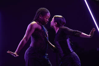 Two dancers stand close together in a dance pose, gazing into each other's eyes. The image is bathed in an atmospheric purple light.
