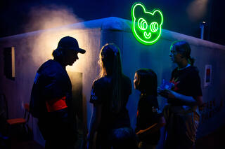 Drei Kindersilhouetten stehen vor einem Gebäude mit einem grünen Bärenkopf als neon Leuchtreklame. Sie stehen an wie bei einem Club.