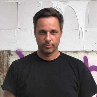 Mann in schwarzem T'Shirt vor einer Graffiti-Wand schaut direkt in die Kamera