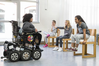 Frau im Rollstuhl unterrichtet in einem Tanzstudio drei Mädchen, die auf Stühelen sitzend, tanzend.