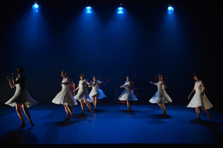 Eine Gruppe Tap-Tänzerinnen in weißen Kleidern auf einer blau beleuchteten Bühne.