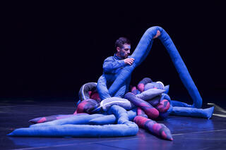 Ein Performer im blauen Kostüm hantiert mit überdimensionalen blauen Würsten aus Stoff.