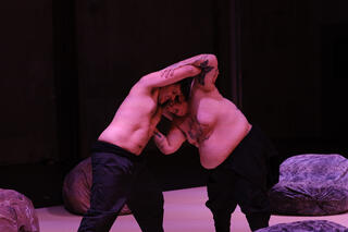 Zwei Tänzer*innen auf der Bühne, mit nackten Oberkörpern, miteinander ringend 