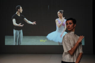 Im Vordergrund eine Person, weiß gekleidet, die einen Arm schräg über die Brust legt und leicht schräg steht. Im Hintergrund direkt hinter der Person eine Ballerina in der gleichen Pose, links davon ein Mann der beide Arme leicht anhebt.