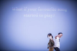 Eine Frau und ein Mann tanzen Rücken an Rücken. Auf einem blauen Hintergrund steht "What would you do if your favourite song started to play?"