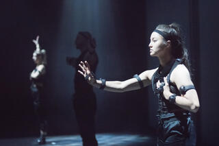 Drei Performer*innen mit Motion-Capture Anzügen stehen nebeneinander auf der Bühne. Sie führen verschiedene Posen aus, indem sie ihre Arme ausstrecken.
