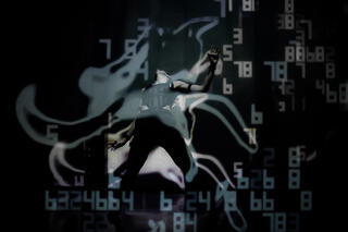 Ein*e Performer*in im Motion-Capture Anzug steht mit dem Körper nach hinten gebeugt auf einer Bühne. Er*sie ist von Projektionen umgeben, die sich aus wellenartigen Formen und Zahlen zusammen setzen.