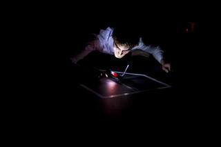 Eine Person liegt in einem dunklen Raum mit dem Bauch auf dem Boden. Das Gesicht der Person wird nur durch ein techisches Gerät erleuchtet, welches sie in der Hand hält.