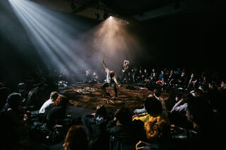 Auf der Bühne, die mit Erde ausgelegt ist, stehen zwei Performer*innen in expressiven Posen in der Mitte einer Menschenmenge.