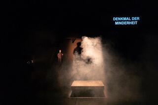 Ein*e Performer*in steht im Nebel hinter einem Tisch. Rechts oben steht "Denkmal der Minderheit2