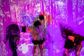 Drei Tänzer*innen bewegen sich stürmisch stehend vor einem lila bunten Hintergrund.