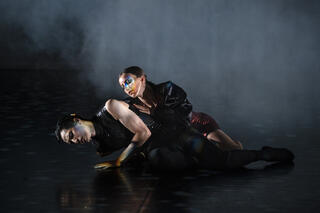 Zwei Tänzer*innen in dunkler Kleidung auf dunkler Bühne. Ihre Gesichter sind in buntes Licht getaucht 